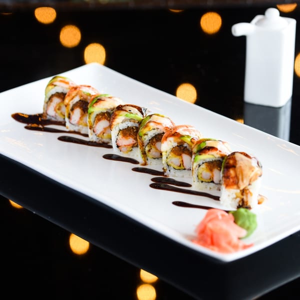 A white rectangular plate holds a row of shrimp tempura sushi rolls drizzled with sauce.